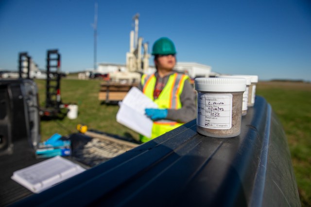 A close up of soil samples in plastic containers in the foreground. Out of focus in the background is a person in a bright yellow vest and hard hat holding a notebook.