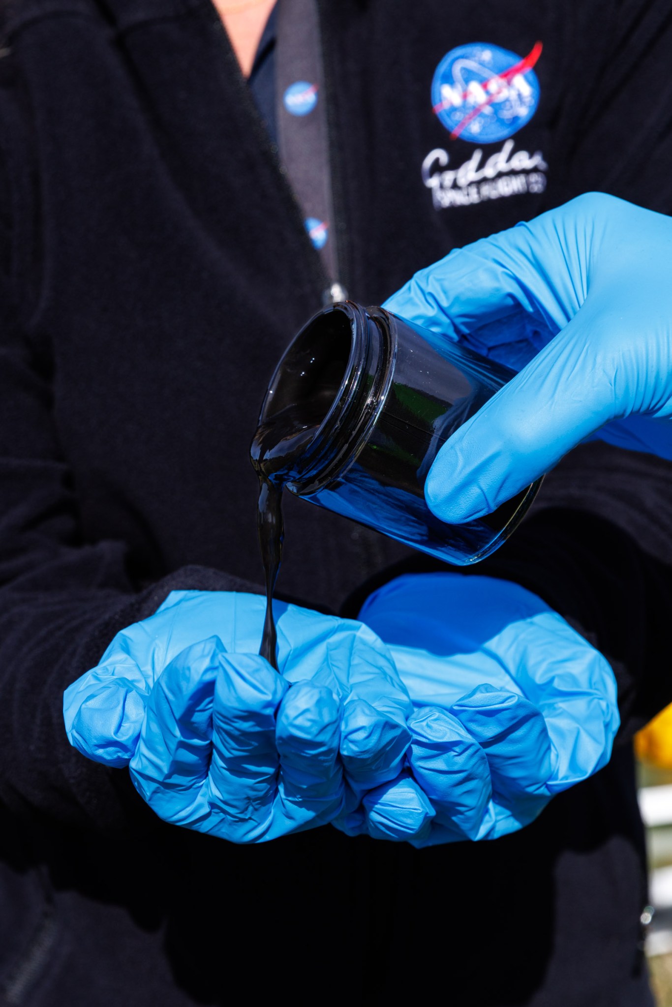 Two hands with bright blue gloves cupped together to catch a thick, black liquid being poured from a container.