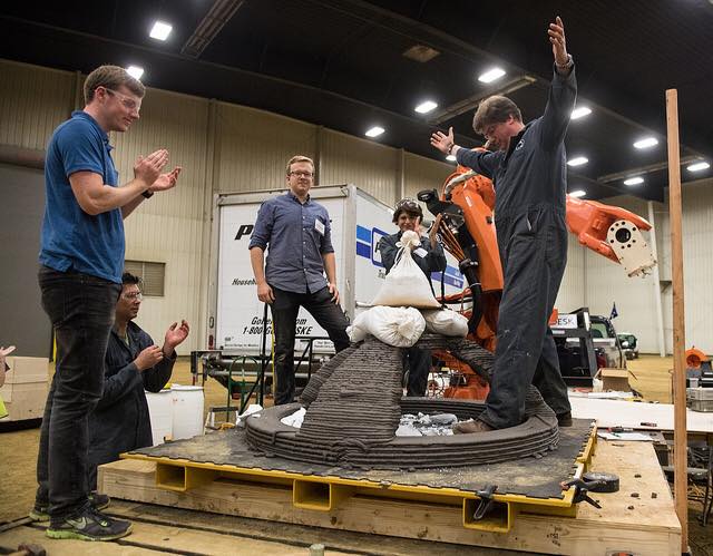 Four men and one woman surround a 3D-printed dome. The dome is being durability-tested using sandbags and the weight of one of the men, who is dressed in a gray work jumpsuit and is raising his arms in victory. The other four team members clap and look happy with the results of the durability test. The team is inside a warehouse-like structure with high ceilings. A white and blue Penske cargo truck is parked behind the team. Next to the team is an orange mechanical arm.