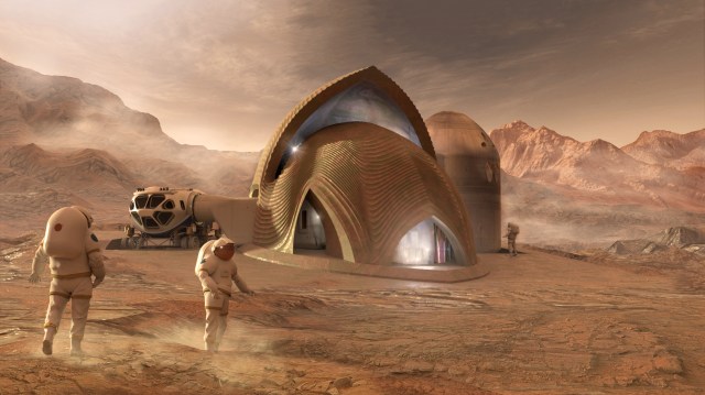 This is a digitally generated image mockup that shows three people outside a 3D-printed habitat on what is meant to look like the surface of Mars. The three subjects are wearing space suits and are walking around the rocky terrain with the habitat in the center of the image. The habitat is angular, with triangular openings and a vehicle affixed to a hatch on the left side. Rocky mountains are in the background, set against an orange-ish, dusty sky.