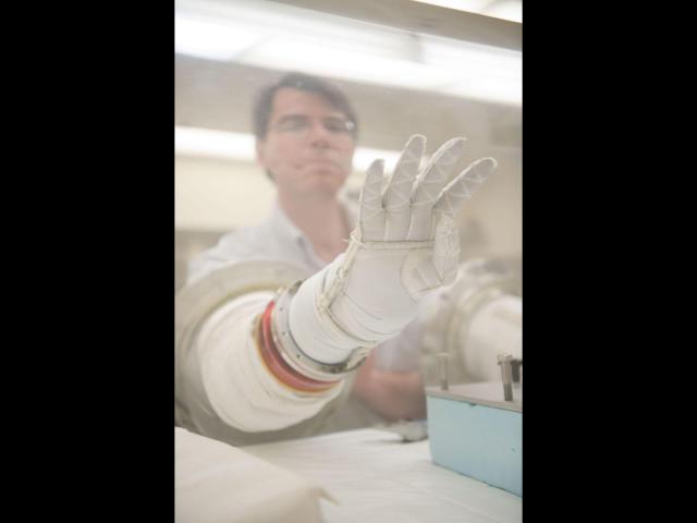In the foreground, there is a white glove that hovers inside a clear vacuum chamber. In the background, slightly out of focus, there is a light-skinned man with short, dark hair who is wearing clear protective glasses as his arm maneuvers the glove from outside the chamber. Behind the man is a blurry view of fluorescent lighting on the ceiling of a brightly-lit room.