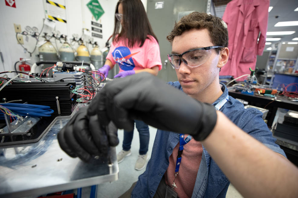 Student with black gloves does work on a piece of equipment in a lab.