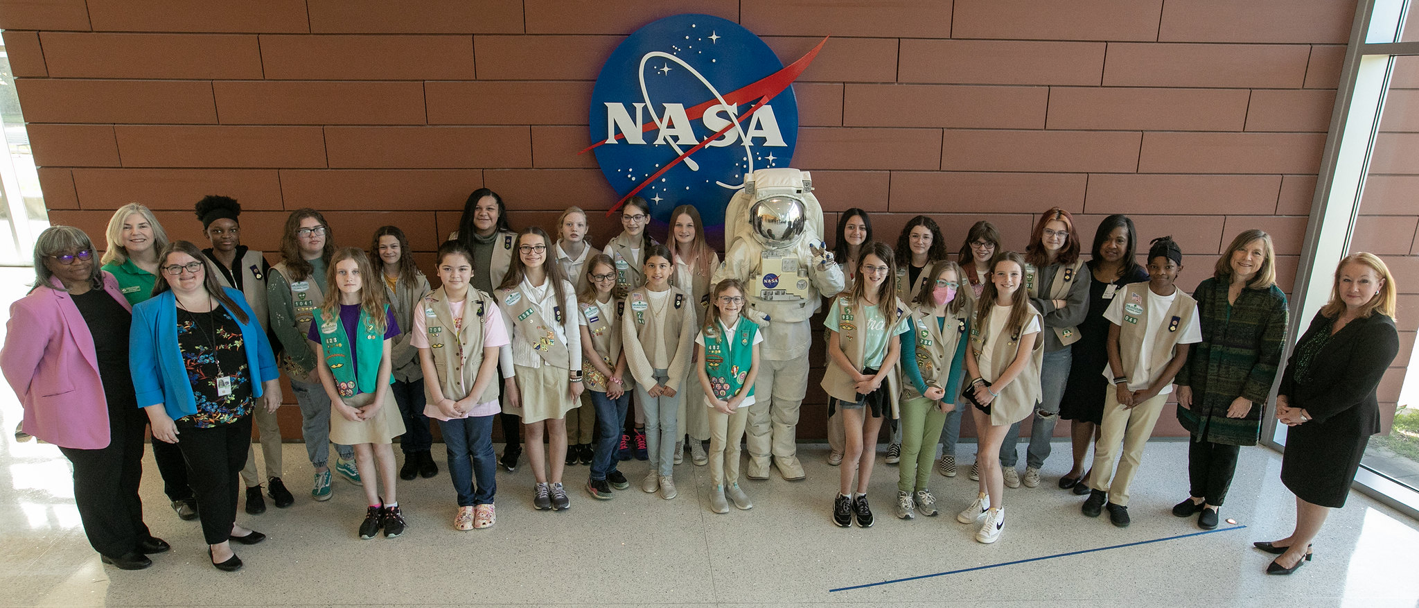 A group of girl scouts poses with a NASA Langley public relations specialist in an astronaut suit in front of a NASA sign.