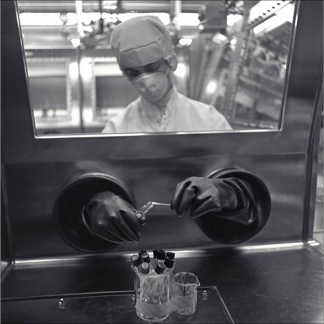 A black and white photo of NASA biologist Elaine Muñoz, wearing clean room attire, handling a lunar sample through one of the lab's glove boxes.