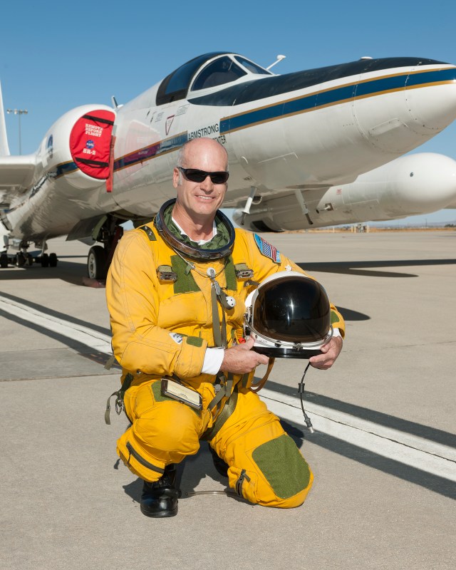 Portrait of a pilot in an orange space suit kneeling down to the ground and holding a helmet in his hands. The ER-2 aircraft can be seen in the background.