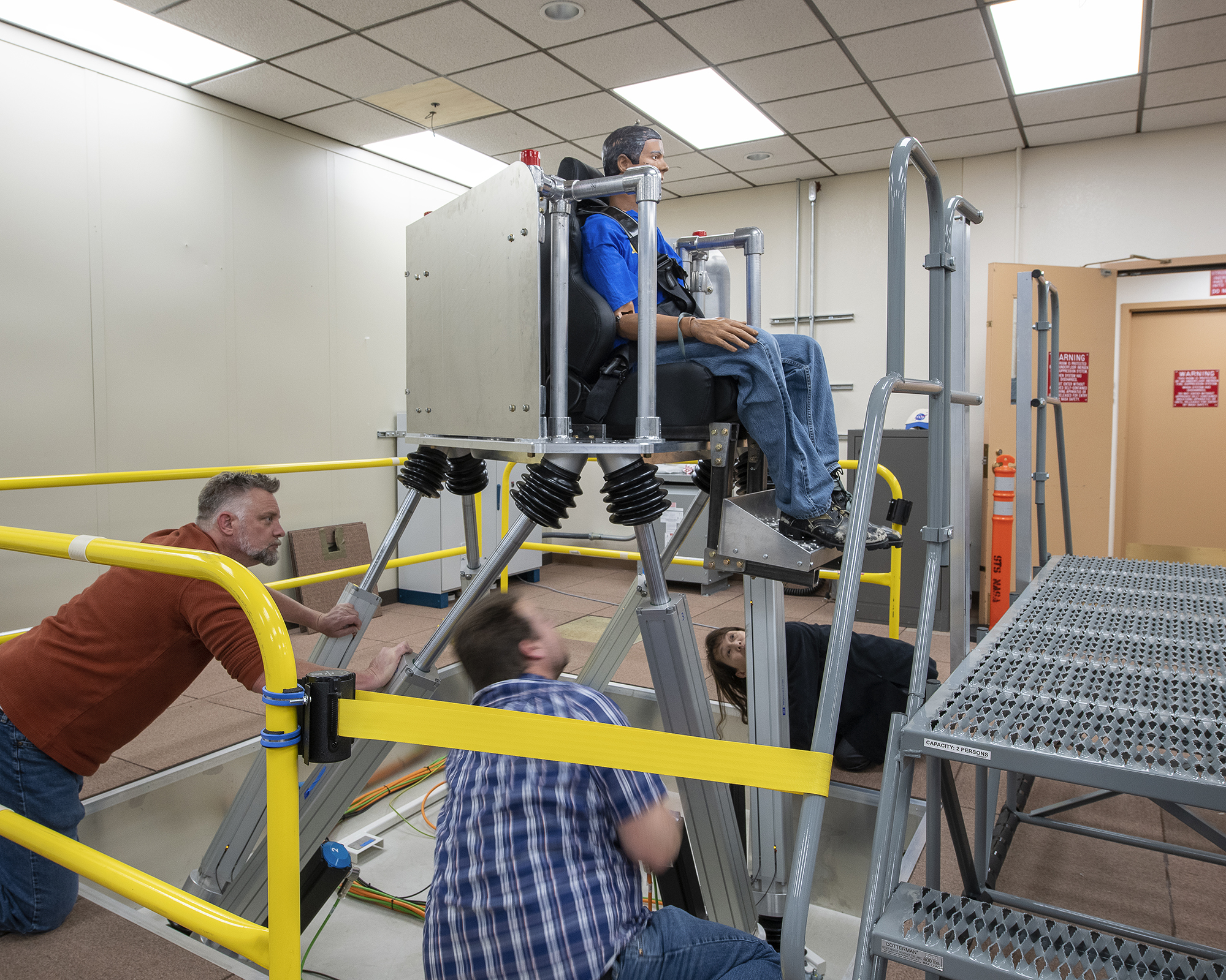 NASA researchers Jeanette Le, Curt Hanson, and Erik Waite, look on as pieces of a newly installed motion simulator are tested at NASA Armstrong Flight Research Center in Edwards, California on April 26, 2023. The simulator includes virtual reality goggles depicting an aircraft cabin and city environment, as well as noise and seat motion, to simulate an air taxi ride.