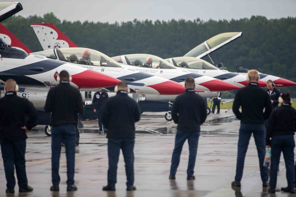 The Thunderbirds flight team stands with their backs to the camera. The Thunderbird F-16 jets lined up in a row on the tarmac at Wallops Flight Facility's airfield. The Thunderbirds are mostly white, with a red tip and blues stripes.