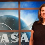 A photograph of a woman with shoulder-length brown hair smiling. Behind her, the NASA insignia is displayed.