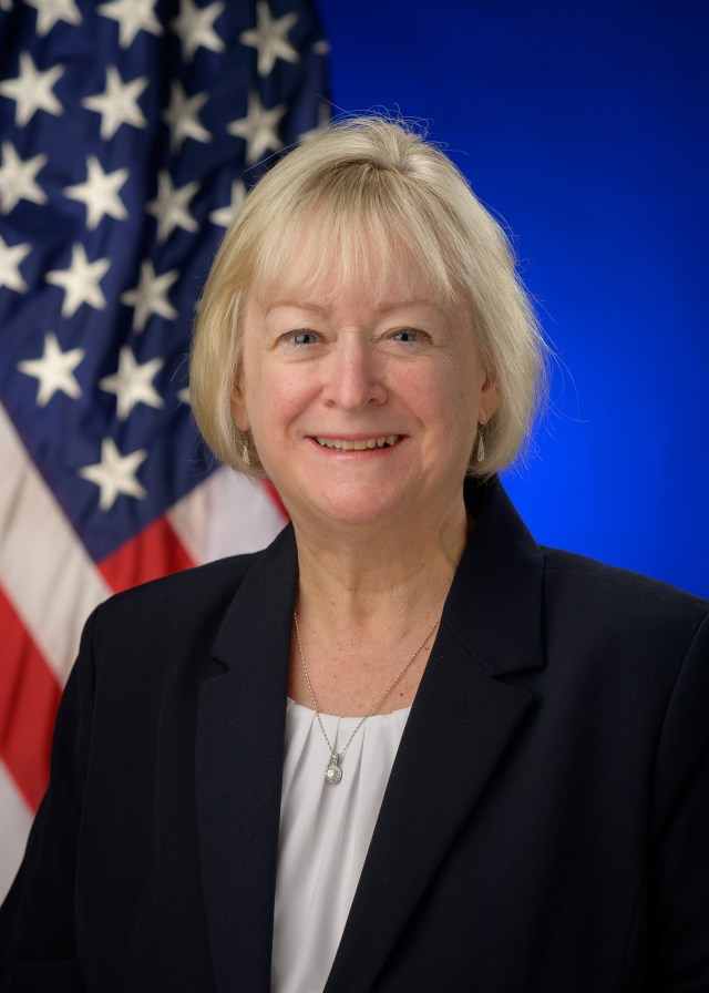 Carol Carrol wearing a dark suit and white top. The U.S. flag behind her against a blue background.