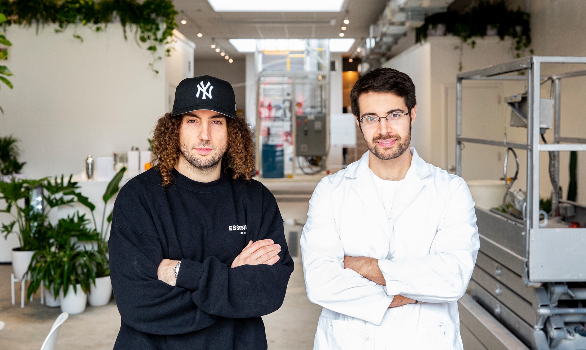 Two fair-skinned men stand facing the camera with their arms crossed over their chests. The man on the left is wearing a dark sweatshirt with a white text label on the left breast. He has shoulder-length curly brown hair and is wearing a navy New York Yankees baseball cap. The man on the right is wearing a white button-down shirt and a white undershirt. He has short, dark brown hair and is wearing wire-rimmed glasses. They stand in front of a laboratory-style workspace, with many plants and machines in the background.