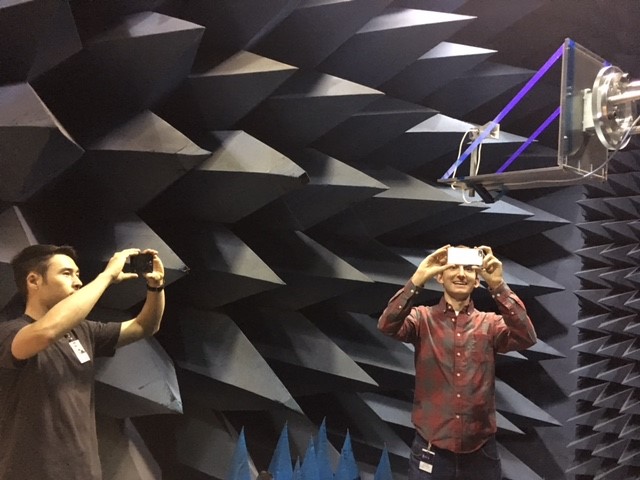 Two men stand in a room with spikes on the walls. They are each holding up a cell phone to take photos of a satellite antenna, which is above them in the top-right corner of the frame. One man is wearing a red button-down shirt and is smiling. The other, in a dark t-shirt, has a badge on his lapel.