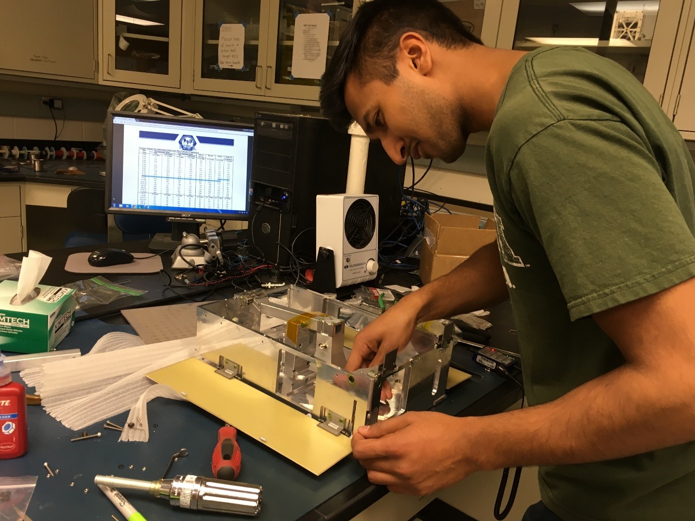 A light-skinned man leans over a CubeSat prototype as he works to assemble the device. His workstation is messily arranged with screws, screwdrivers, a marker, foam sheets, a desktop computer with data displayed on the screen, delicate task wipes, and other items commonly found in a science lab. The man is wearing an olive-green t-shirt and is focused on the prototype. In the background is a counter with other lab items on top of it, as well as white cabinets with supplies inside.