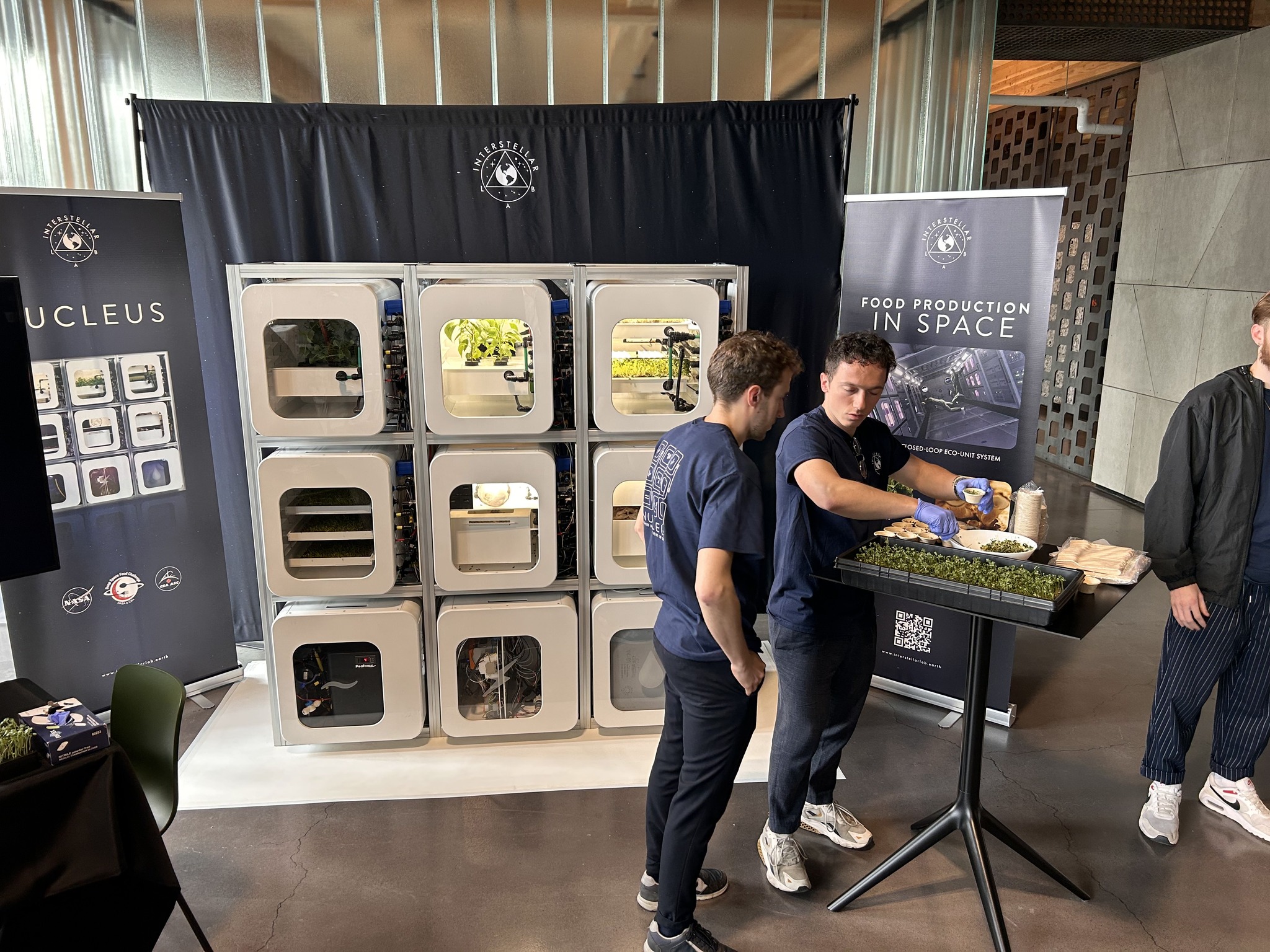 Two white men with brunette hair, wearing navy blue t-shirts and black pants with sneakers, stand in front of a food system demonstration station comprised of nine incubator cubes with plants/vegetation inside. One of the men stands in front of a black table and, while wearing light blue gloves, spoons alfalfa sprouts from a large bowl into a small sample cup. Also on the table are wooden spoons, more sample cups, and a tray of alfalfa sprouts. Behind the demonstration station are three navy backdrops, which include affiliated logos and graphic demonstrations showing how the food system works.