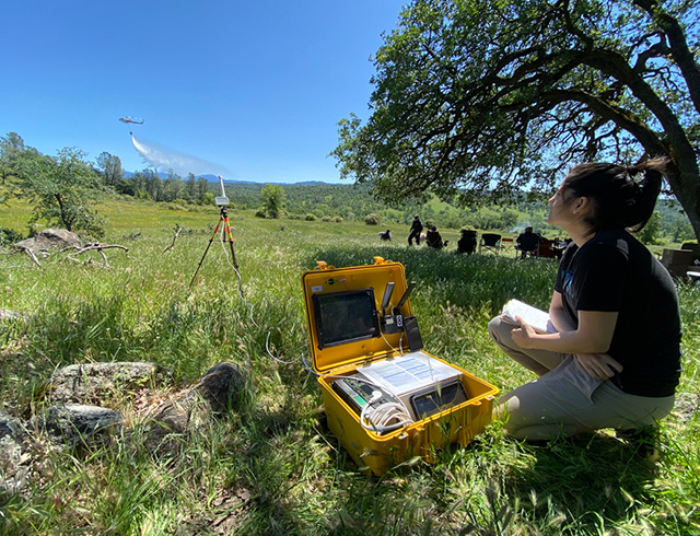 A person kneels in the grass next to a yellow case containing hardware, while a helicopter drops water in the distance