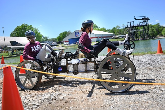 Two students pedal on their rover on the gravel course with water and a helicopter display in the background.