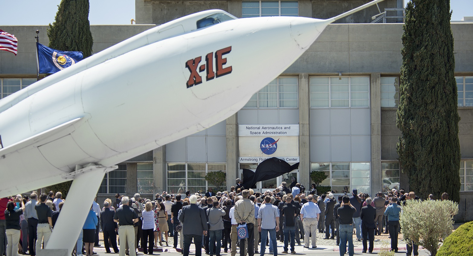 Group of people stand in front of a NASA Armstrong building, facing the building. A black covering is pulled down, revealing a new sign on the front a the building.
