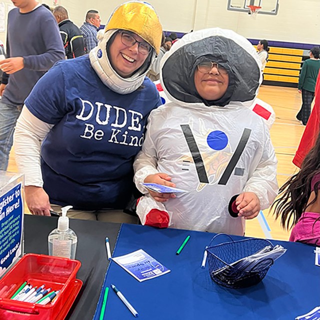A woman wearing a plastic space helmet and a young student wearing an inflatable astronaut costume smile while working on a craft project during an outreach event