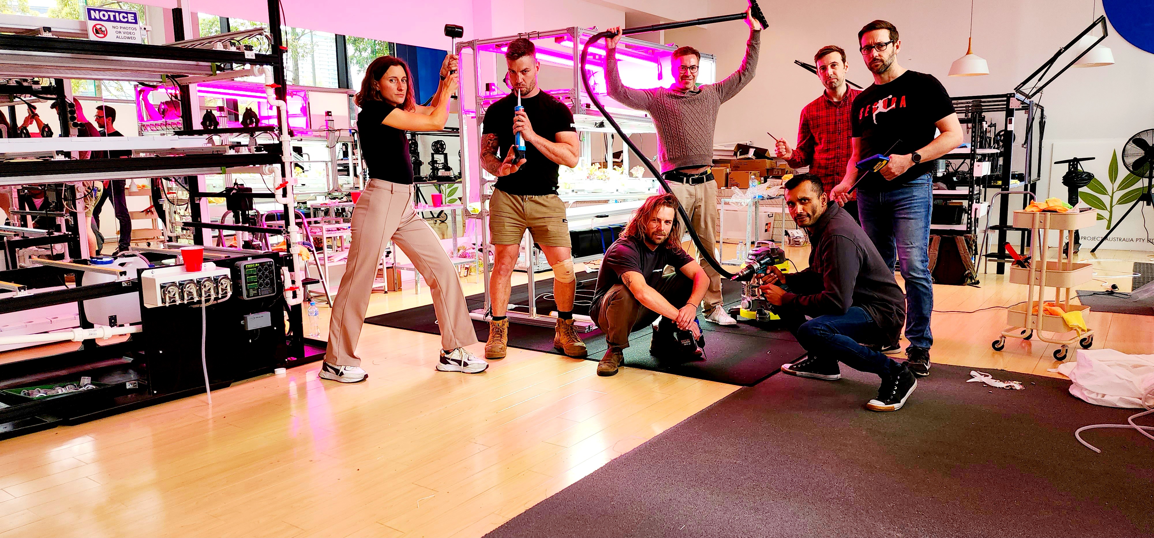 Seven adults stand in various poses in a room full of technological equipment, crop incubators with magenta lights, and research stations. From left to right: a dark-haired woman stands in a wide, twisted stance and holds a rubber mallet in the air; a man with short dark hair and tattoos on his right arm stands facing forward and playfully holds a caulk gun to his mouth; a man with long dark hair and a beard squats down low and holds a drill; a man with short dark hair and glasses stands in a wide stance and holds the hose of a shop vacuum over his head; a man with short dark hair and a beard kneels down low and holds a drill; a man with short dark hair stands slightly behind the group and holds a small screwdriver-like object; a man with short dark hair, a beard, and glasses stands in a natural pose and holds a temperature gun in his left hand and a thin pole in his right hand.