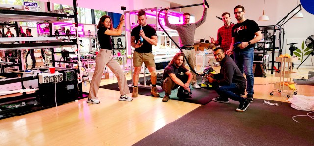 Seven adults stand in various poses in a room full of technological equipment, crop incubators with magenta lights, and research stations. From left to right: a dark-haired woman stands in a wide, twisted stance and holds a rubber mallet in the air; a man with short dark hair and tattoos on his right arm stands facing forward and playfully holds a caulk gun to his mouth; a man with long dark hair and a beard squats down low and holds a drill; a man with short dark hair and glasses stands in a wide stance and holds the hose of a shop vacuum over his head; a man with short dark hair and a beard kneels down low and holds a drill; a man with short dark hair stands slightly behind the group and holds a small screwdriver-like object; a man with short dark hair, a beard, and glasses stands in a natural pose and holds a temperature gun in his left hand and a thin pole in his right hand.
