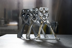 Three organic-looking structures of gray metal stand on a smooth table with a blurred, laboratory background. The pieces look identical, but they range from shiny silver metal in the front to matte gray metal in the back.