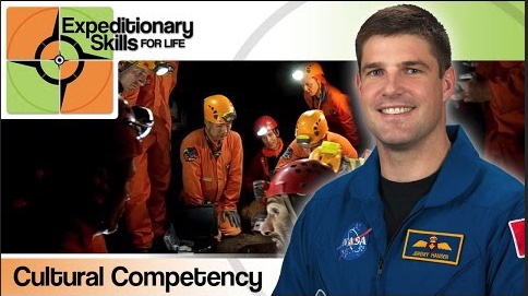 A man in a blue astronaut jumpsuit with the words - Expeditionary Skils, Cultural Competency with people in orange jumpsuits and helmets in the background