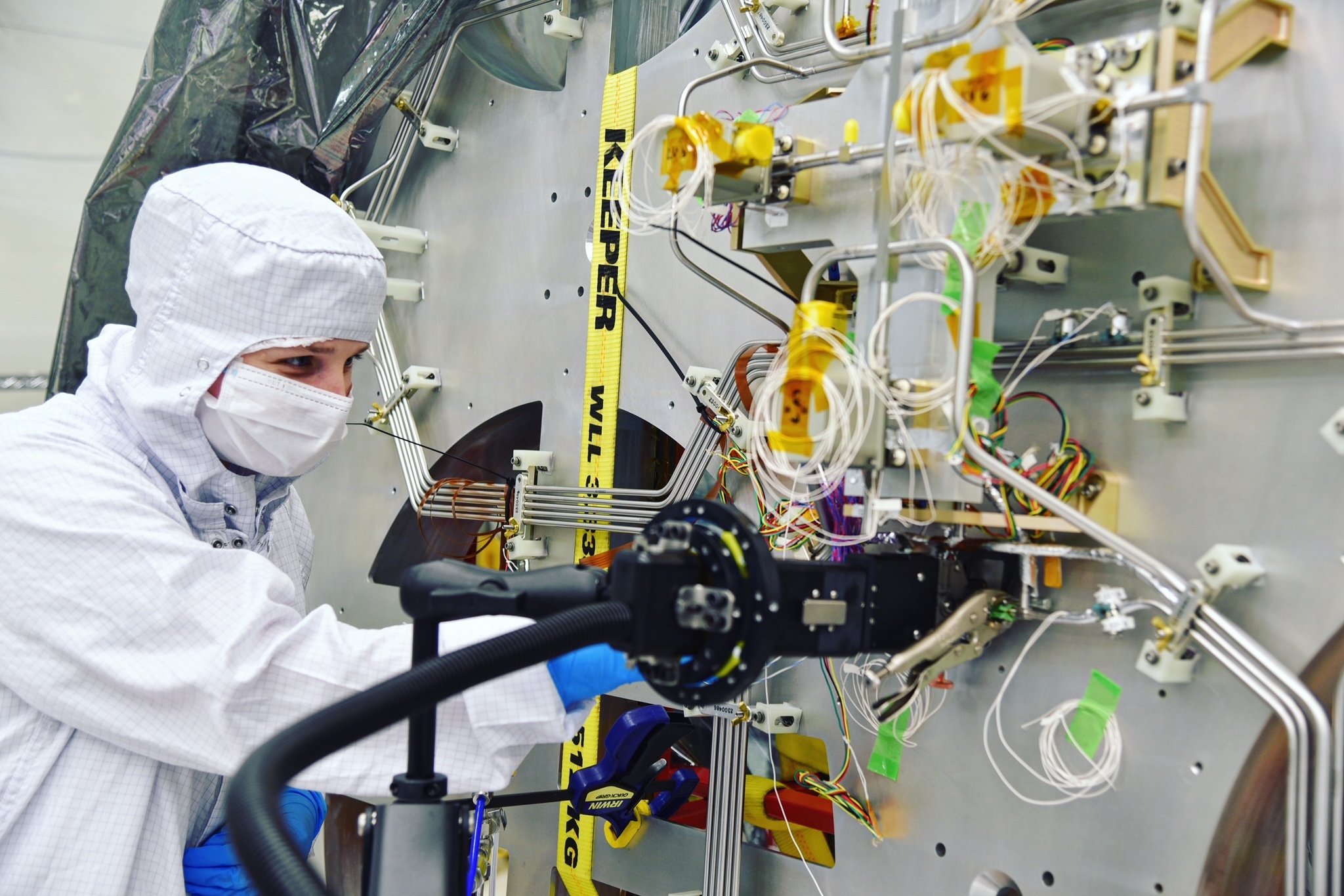 A woman in a white suit, white mask, and blue gloves leans in to weld against a background of hardware with protruding wires.