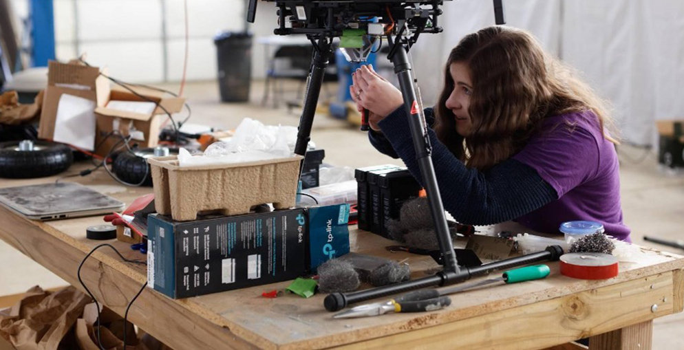 A researcher works to assemble a small spacecraft technology payload perched on a wooden tabletop in a ground-based technology facility.