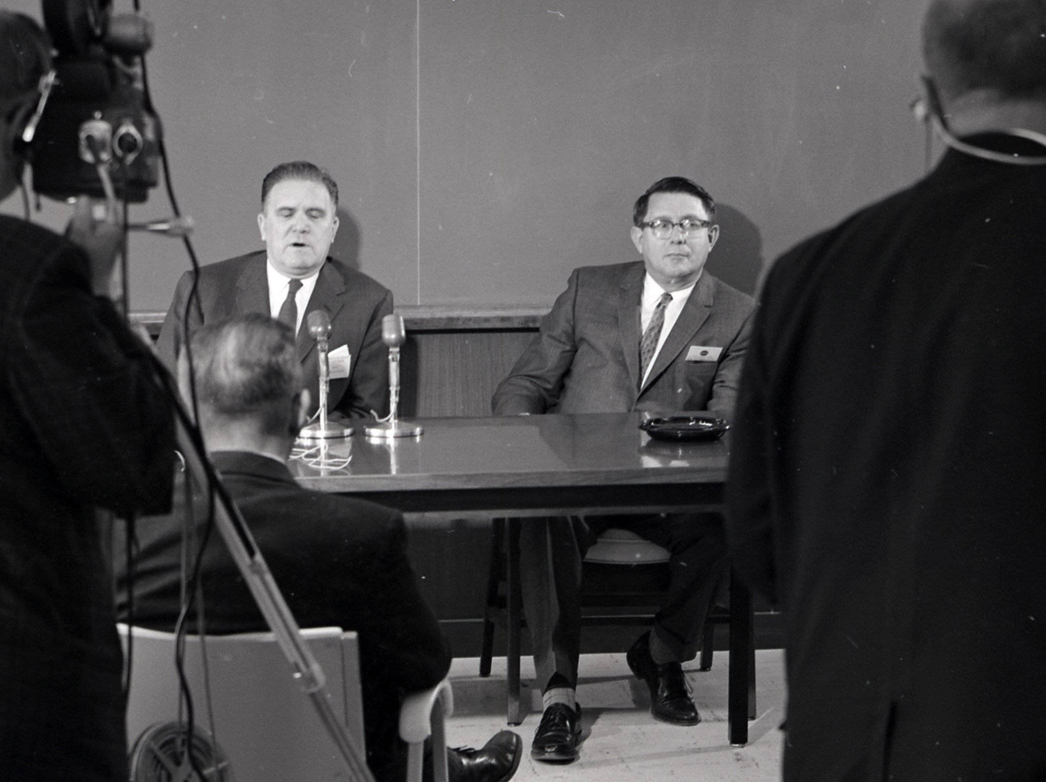 Two men at desk facing cameras.