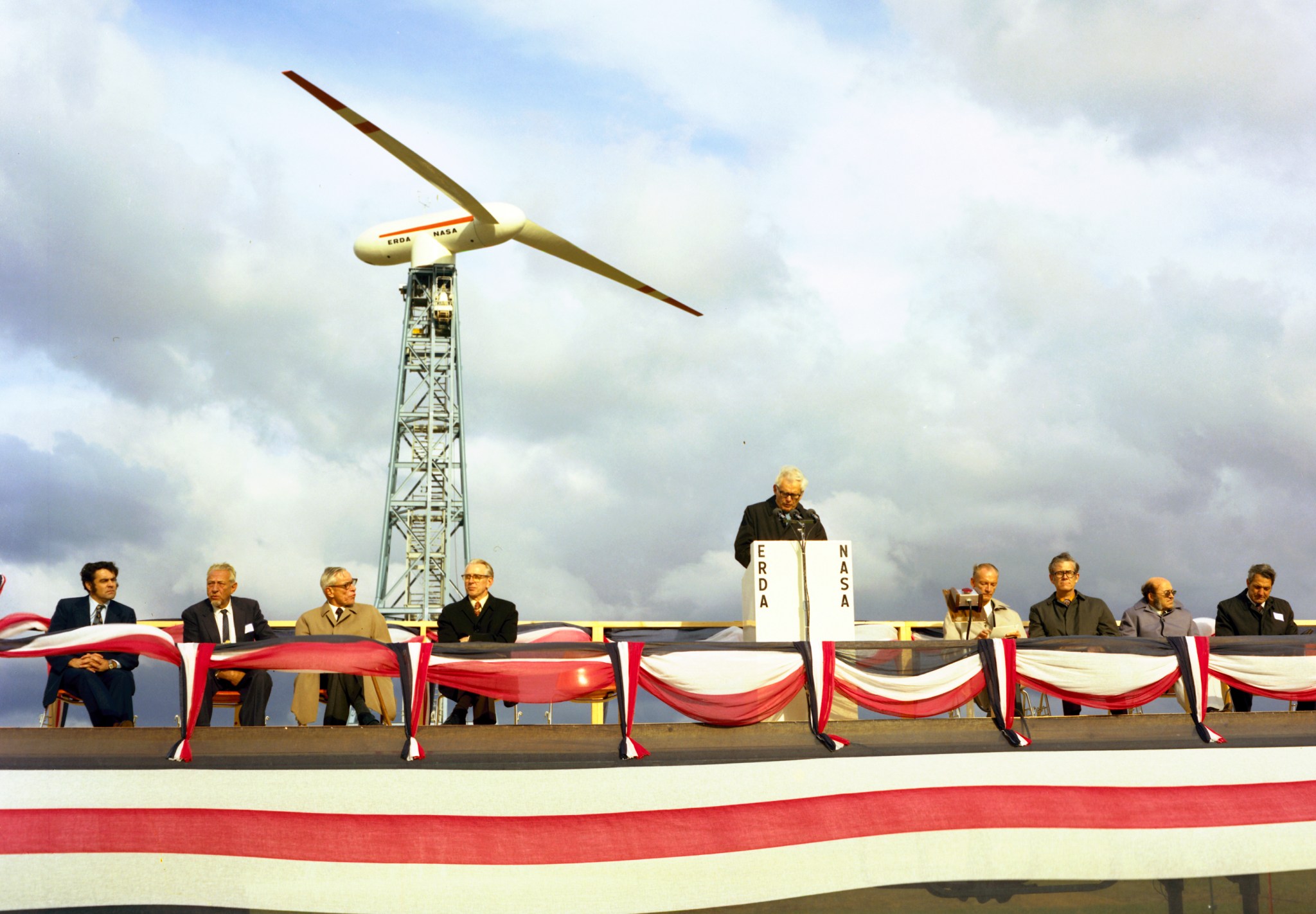 Group of men on podium in front of wind turbine.