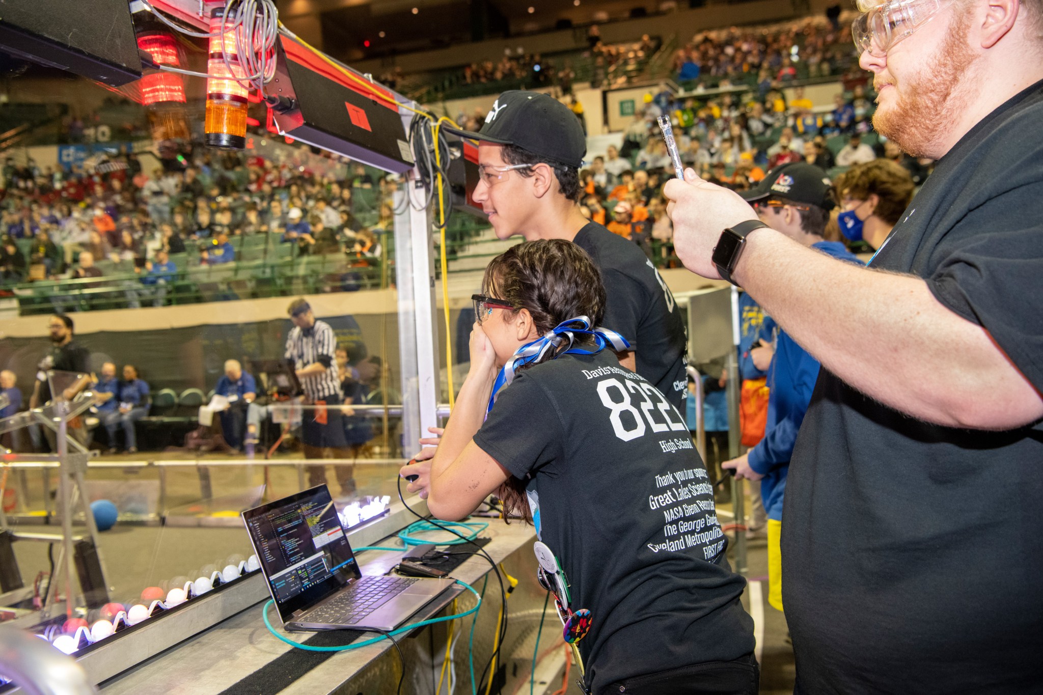Students behind glass are using a computer and controlling robots in the competition area during the First Robotics competition.