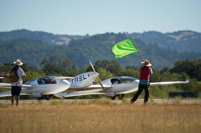 With green mountains and a clear blue sky in the background, a man stands with his back to the camera as he waves a lime green flag. He is wearing a light blue shirt with long pants, a red vest, and a wide-brimmed hat. Another man, wearing dark shorts, a white t-shirt, a dark backpack, and a wide-brimmed hat stands off to the left. In front of the men is a large white aircraft with a center propeller and two cockpits on either side. Part of the aircraft’s name (Pipistrel-USA) is visible on the side in all-caps, with the NASA meatball logo near the center propeller.