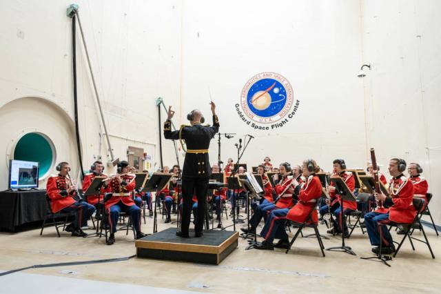 A conductor with back to the camera holds up his baton in front of two-dozen musicians dressed in red. A blue NASA Goddard logo appears on the wall behind them.