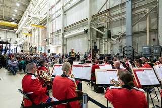 Red-garbed musicians and their music appear in the foreground of the photo, with a crowd of seated and standing onlookers in the background, all situated within a metal and concrete building with exposed support structures and gantries.