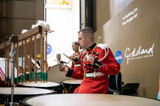 A percussionist in red uniform plays timpani in front of a brown metal door with the NASA logo and the word "Goddard" written on it.