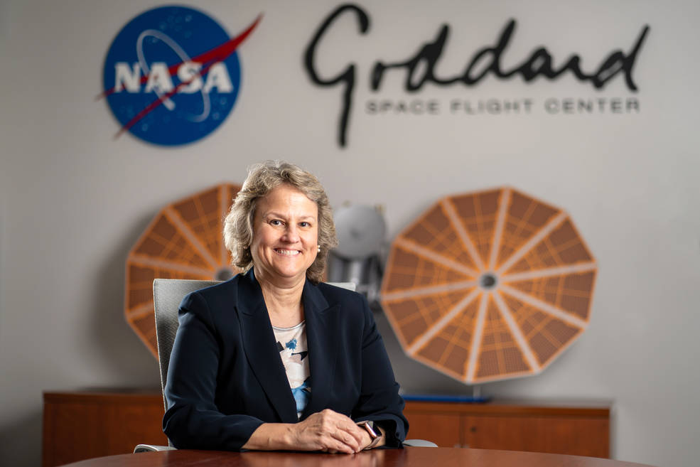DeeDee Healey sits at a table with her hands crossed, posing for a portrait with the NASA and Goddard logos behind her. Two solar arrays from a model of the Lucy spacecraft sit on a table behind her.