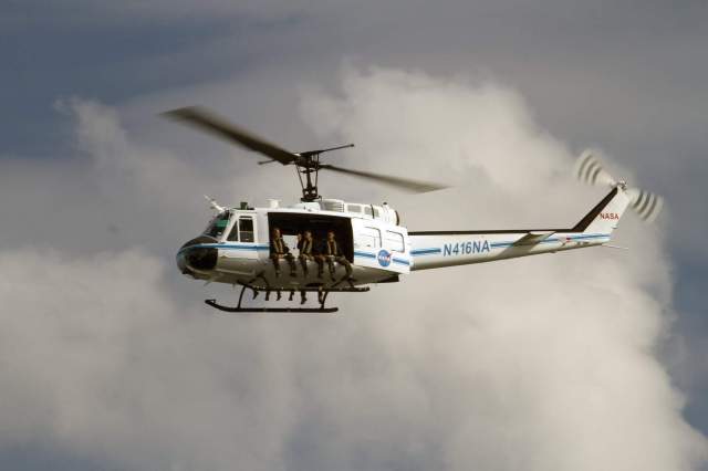 Image of a helicopter in flight with a large cloud in the background and 3 people sitting on the edge of the helicopter.