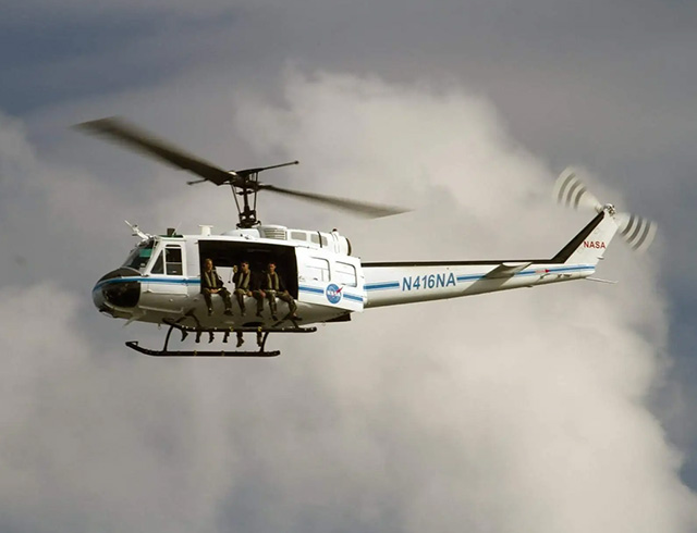 Image of a helicopter in flight with a large cloud in the background and 3 people sitting on the edge of the helicopter.