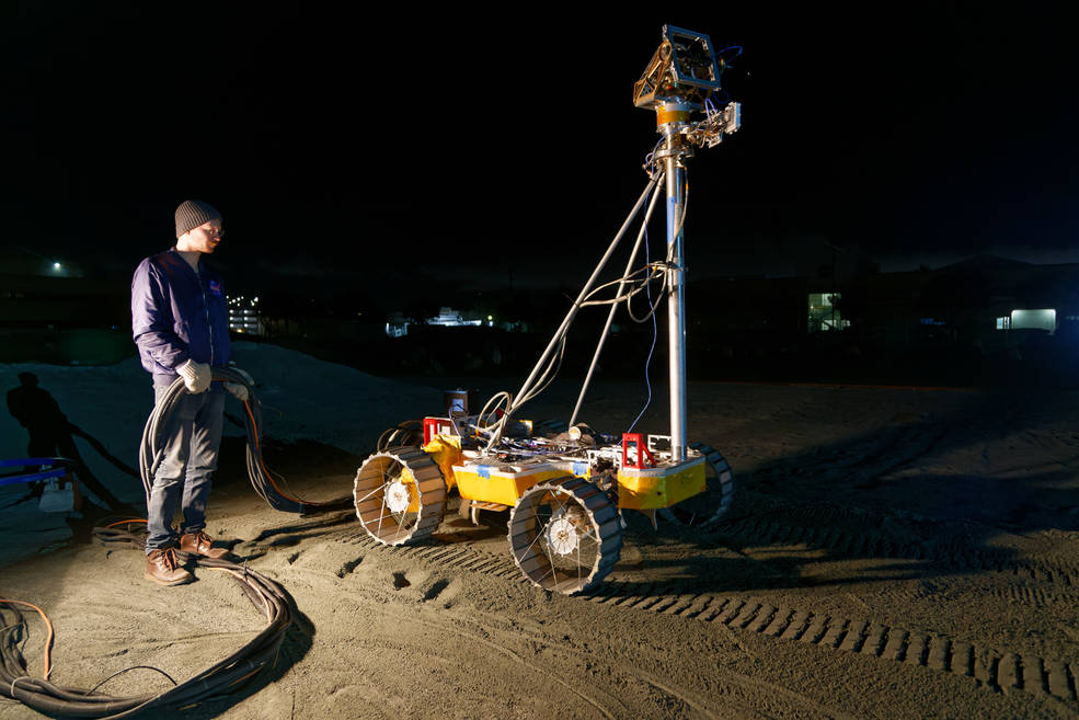 An engineer stands beside the latest prototype of the VIPER (Volatiles Investigating Polar Exploration Rover), known as MGRU3 (Moon Gravitation Representative Unit 3) during a night time test in the Roverscape at NASA's Ames Research Center.