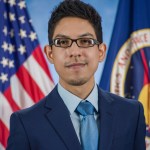 A portrait photograph of a man with short black hair and glasses looking at the camera. Behind him are the United States flag and a flag with the NASA seal.