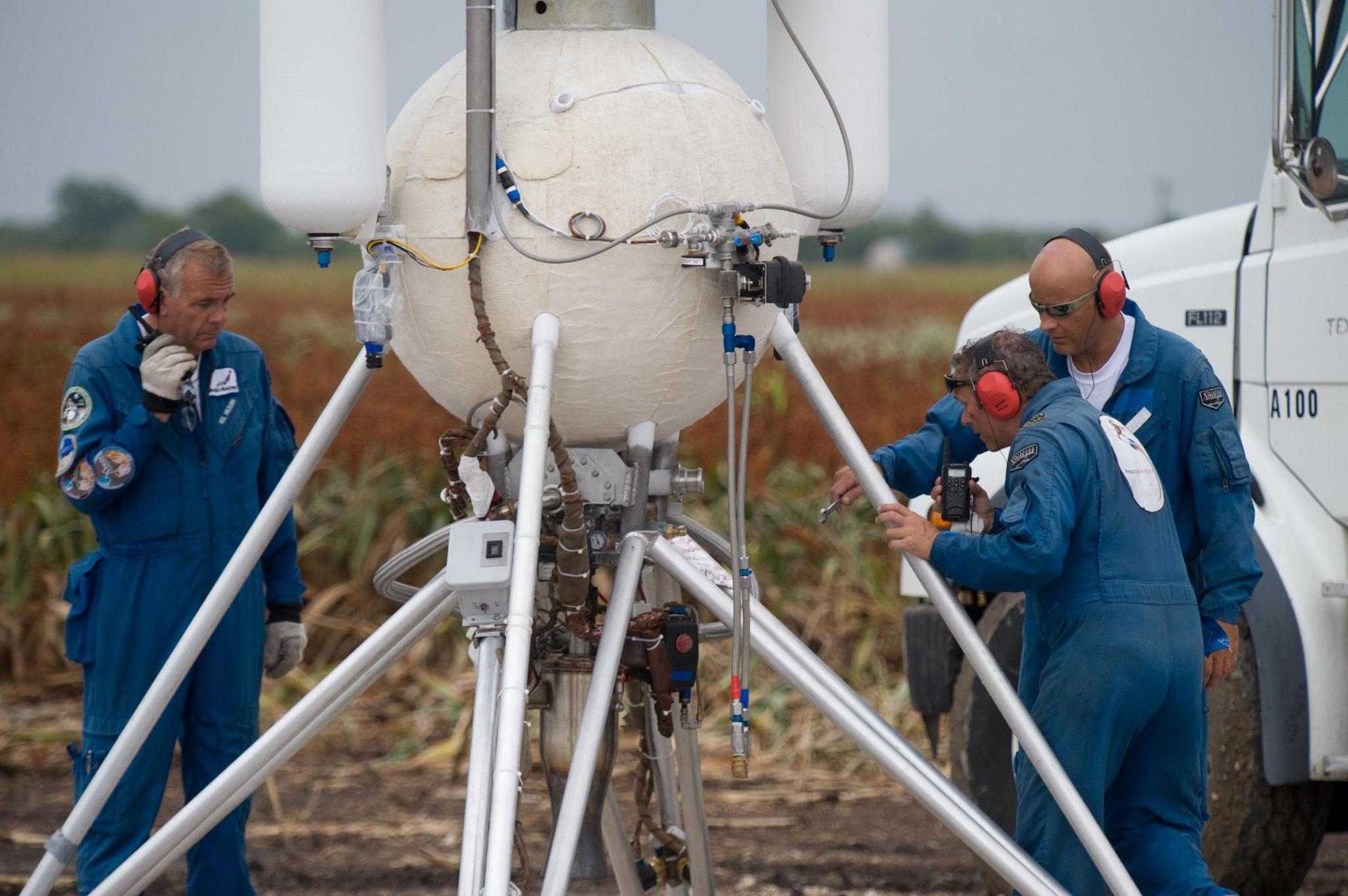Three light-skinned men stand around a white rocket-like structure, which features a spherical object atop a four-legged metal stand. There are fuel tanks, valves, wires, and other mechanisms all over the body and base of the rocket. The men are each wearing blue jumpsuits and black and red safety headphones. Behind the group, out of focus, is a corn field and overcast sky.