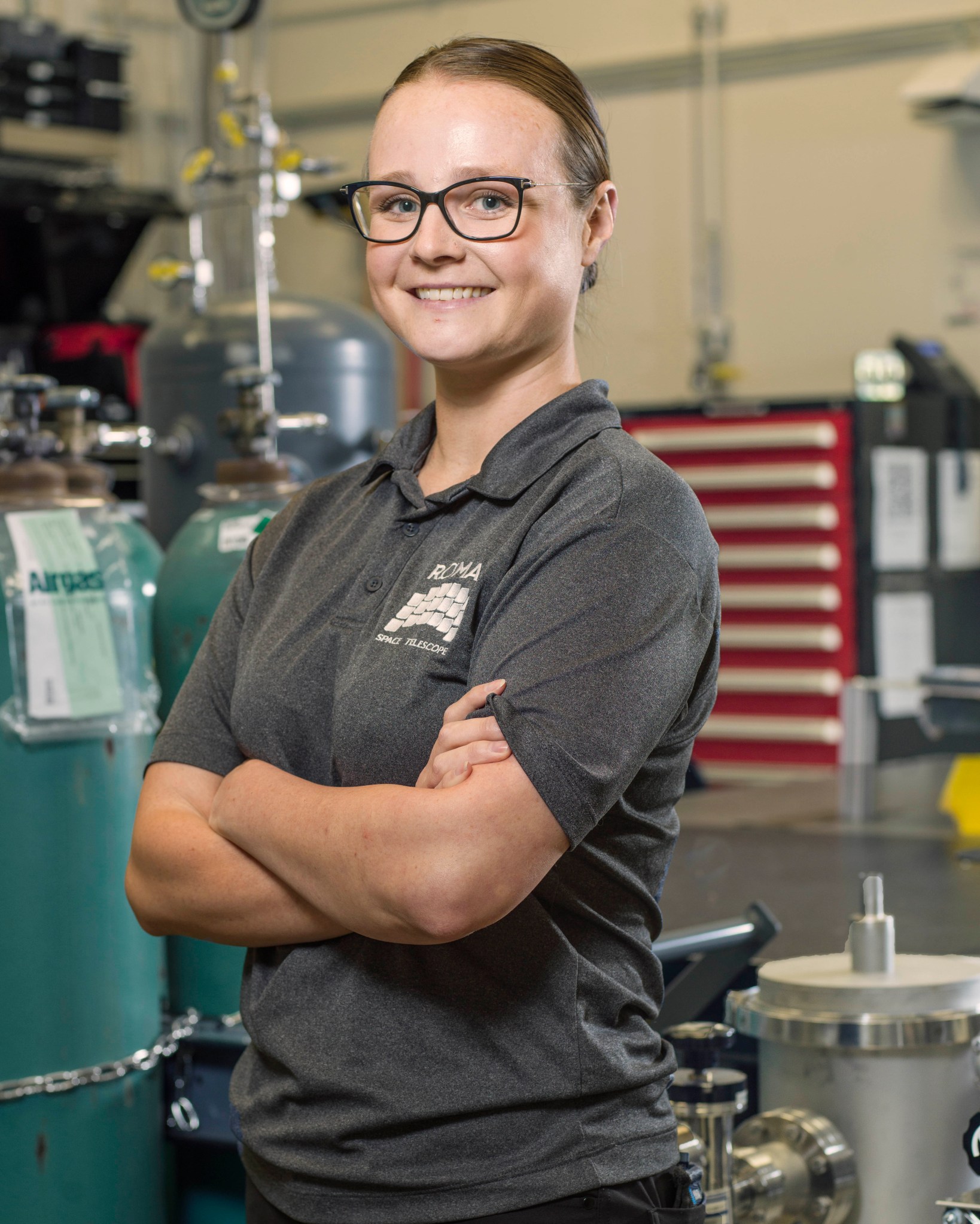 A woman wearing glasses and a collared shirt with the 18-paneled Roman Space Telescope logo stands arms-crossed in front of two green gas tanks in a lab setting.