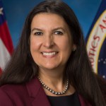 A portrait photograph of a woman with long, dark hair smiling and wearing a maroon blazer and necklace.