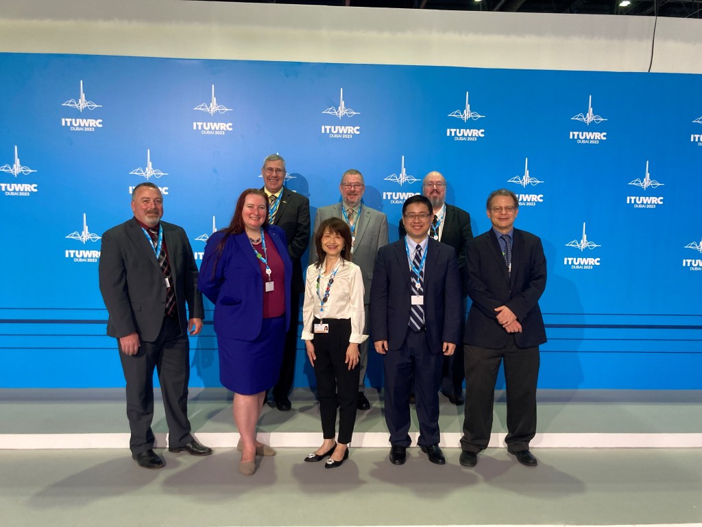 Eight of the NASA members of the U.S. delegation to the 2023 World Radio Conference pose together in front of the conference’s official backdrop, which combines wave forms reminiscent of modulated analog radio waves to form the white logo for Dubai 2023 against a light blue background.