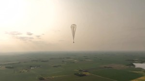 Aerostar’s high-altitude balloon is shown floating above expansive green fields and a small body of water appears in the lower left corner. The sun, low in the sky and out of frame, is casting sunlight through clouds and haze.