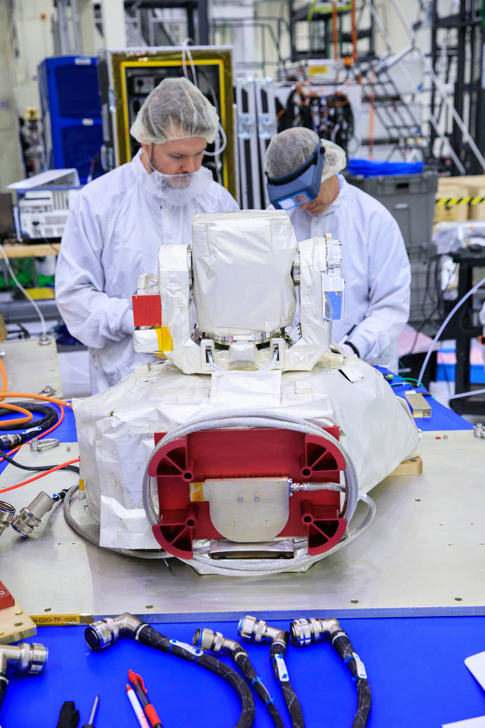 The Orion Artemis II Optical Communications System (O2O) at Kennedy Space Center. In this picture, the payload is in a cleanroom, on a table and is being examined by engineers in white bunny suits.