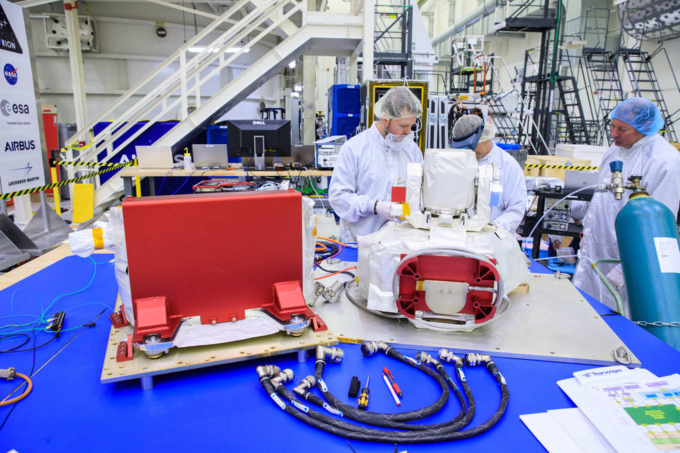 The Orion Artemis II Optical Communications System (O2O) at Kennedy Space Center. In this picture, the payload is in a cleanroom, on a table and is being examined by engineers in white bunny suits.
