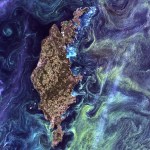 A brown and green island, centered in the middle of the image, surrounded by a dark blue ocean with wispy swirls of greens and teals that illustrate blooms of phytoplankton.