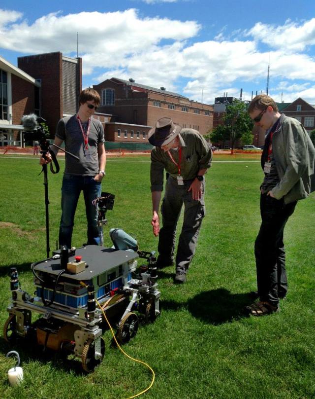 Three light-skinned men stand in front of a robot rover outside on a grassy lawn. A blue, partly cloudy sky is in the background, with several brick buildings in sight. The men are wearing red lanyards with white badges. The center man is slightly bending forward with his hand extended toward the robot. The robot is on six wheels with several wires and buttons affixed to it. The man on the left is holding a camera with a wind shield on a monopod.