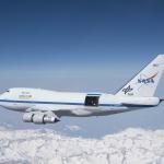NASA's SOFIA jet in the air, with the observatory's door open. The landscape below shows snow-covered mountains.