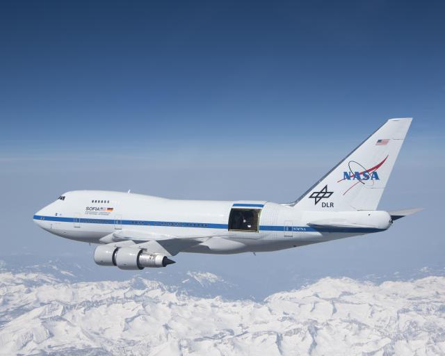 NASA's SOFIA jet in the air, with the observatory's door open. The landscape below shows snow-covered mountains.